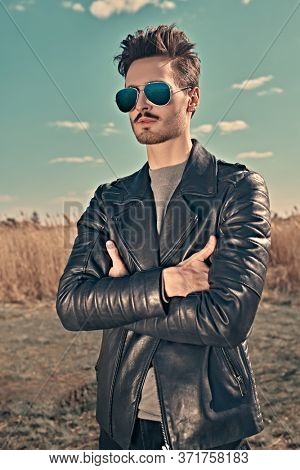 Handsome young man in a black leather jacket and sunglasses stands in the park on the background of reeds. The spirit of freedom and adventure. Men's fashion. 