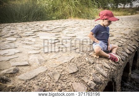 Little Boy Sitting Over Bridge Observing How The Water Flows. Discovering Nature For Children Concep