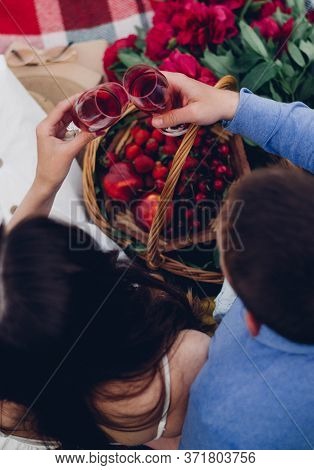 top view of a guy and a girl who are sitting on a plaid blanket and holding glasses with red wine in