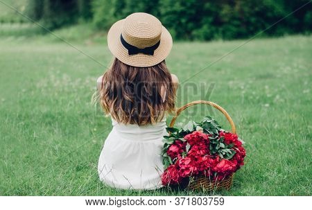 Girl In A White Dress In A Boater Sits With Her Back On The Green Grass Near The Basket With Red Peo