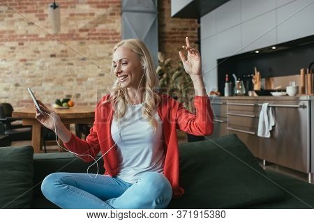 Woman Listening Music In Earphones While Sitting With Raised Hand And Holding Smartphone
