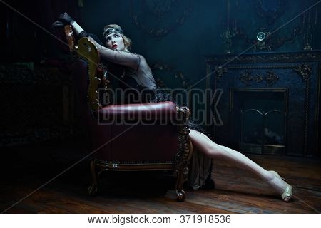 Beautiful woman in an elegant evening dress in an armchair in a rich classic interior. Clothing, makeup and hairstyle in the style of the 1920s.