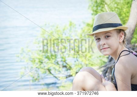 In Summer, On A Bright Sunny Day, A Girl In Hat Ssits Near The Lake.