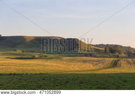 A Grassy Field With Hill With Forests In The Distance. Rural Scene, Country Road And Old Farm In Cou