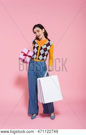 Full Length Of Pleased Woman In Trendy Attire Standing With Gift Box And Shopping Bags On Pink Backg