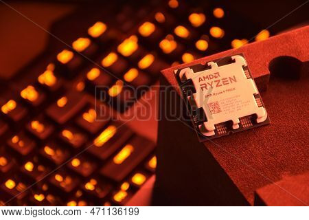 A Close-up Of An Amd Ryzen 9 7900X Processor In A Packaging Box With Backlit Keyboards In The Backgr