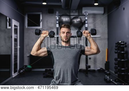 Muscular Guy Lifting Dumbbell While Sitting On A Bench At The Gym. A Young Athlete Using Dumbbells D