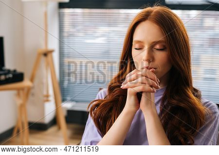 Close Up Portrait Of Sorrow Young Woman Sincerely Praying With Eyes Closed Holding Folded Hands In F