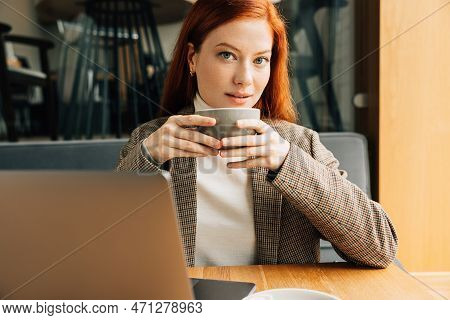 Beautiful Woman With Ginger Hair Looking At The Camera In A Cafe. Businesswoman In A Blazer Holding