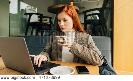 Woman Working As A Digital Nomad In A Cafe. Female Entrepreneur With Ginger Hair Holding A Cup Typin