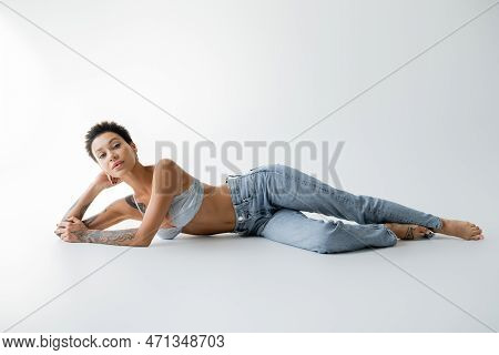 Full Length Of Sexy Brunette Woman In Jeans And Bralette Lying On Grey Background.