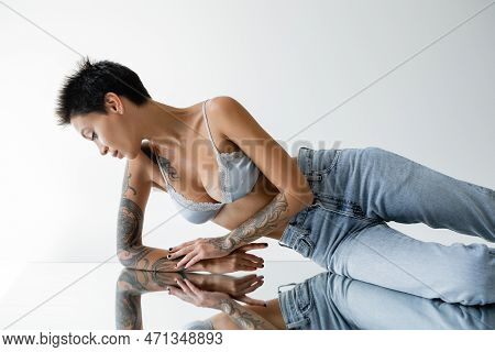 Tattooed Woman In Blue Bralette And Jeans Lying On Mirror On Grey Background.