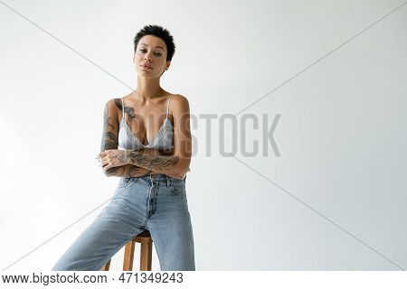 Young Tattooed Woman In Blue Bralette And Jeans Sitting On Stool On Grey Background.