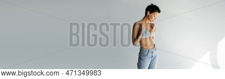 Slender Tattooed Woman In Blue Jeans Touching Straps Of Silk Bralette On Grey Background, Banner.