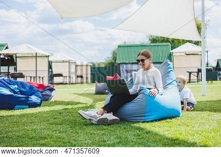 Mother With Laptop, Her Son Playing Outdoors On Green Lawn. Rest And Work With A Child In The Park