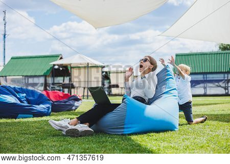 Mother With Laptop, Her Son Playing Outdoors On Green Lawn. Rest And Work With A Child In The Park