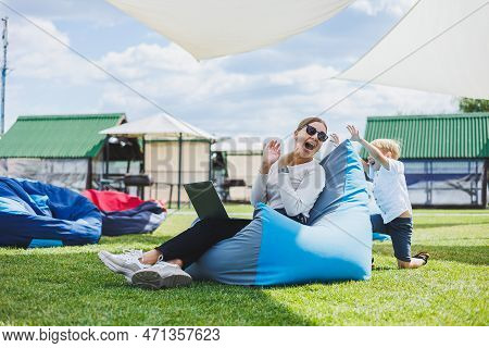 Mother With Laptop, Her Son Playing Outdoors On Green Lawn. Rest And Work With A Child In The Park