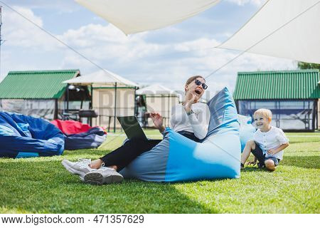 Mother With Laptop, Her Son Playing Outdoors On Green Lawn. Rest And Work With A Child In The Park