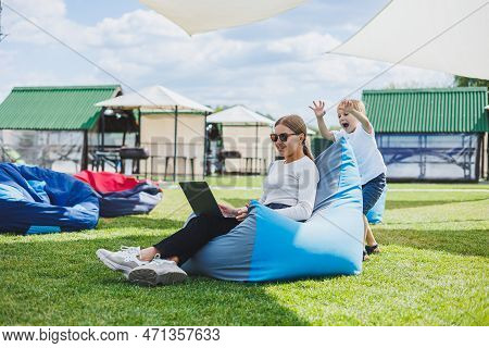 Mother With Laptop, Her Son Playing Outdoors On Green Lawn. Rest And Work With A Child In The Park