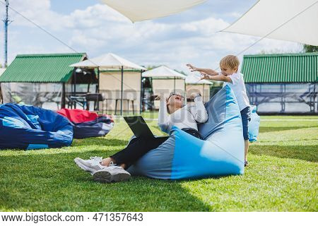 Mother With Laptop, Her Son Playing Outdoors On Green Lawn. Rest And Work With A Child In The Park
