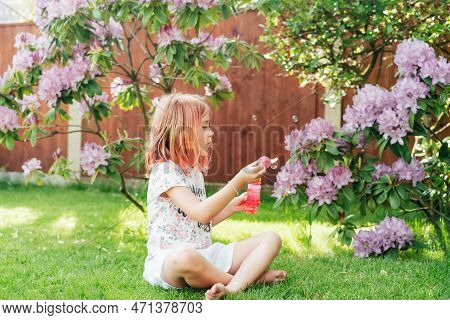 Cute Little Girl With Toned Red Hair Blowing Soap Bubbles While Sitting On A Green Lawn In The Garde