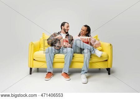 Full Length Of Stylish Multiethnic Men Smiling At Each Other While Sitting With Buckets Of Popcorn O