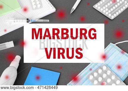 The Word Marburgvirus On Gray Modern Doctor Desk Table Background. Mask, Notepad, Syringe, Sanitizer