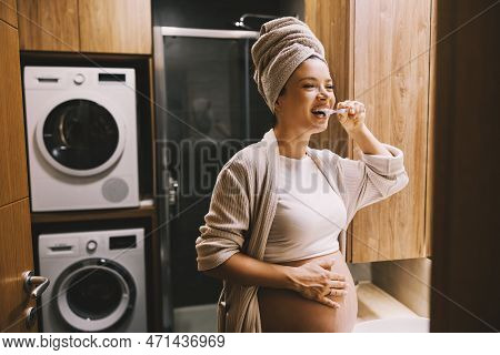 A Happy Pregnant Woman Is Brushing Her Teeth In A Bathroom.