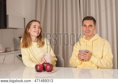 Happy Lovely Couple In Yellow Holding Cups, Eating Red Apples And Sitting By The Table