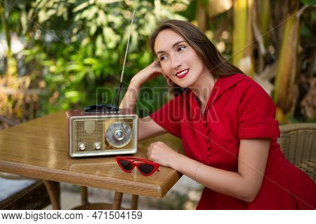 A Beautiful Girl In A Red Retro Style Dress Listens To An Antique Radio