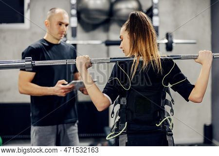 Body Part Of A Woman In A Black Ems Training Suit Standing In The Middle Of The Gym. He Holds A Barb