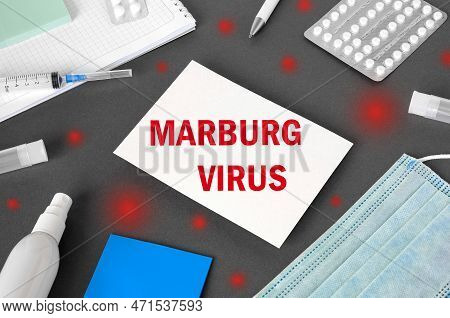 The Word Marburgvirus On Gray Modern Doctor Desk Table Background. Mask, Notepad, Syringe, Sanitizer