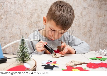Preschooler Boy Burning An Ornament On Wooden Blank With Pyrograph, Sitting At Table At Home. Child 