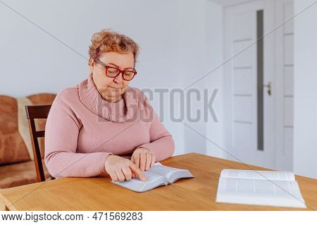 An Older Woman At Home, Glasses Perched On Her Nose, Sits Comfortably In Her Favorite Chair, Reading
