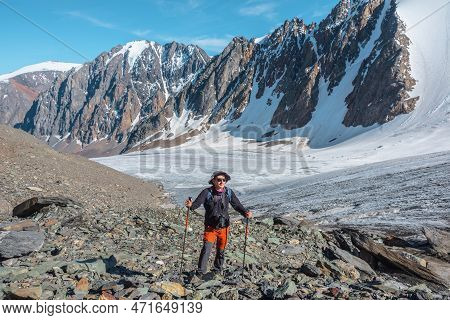 Hiker With Trekking Poles Walks Near Large Glacier Tongue In High Mountains. Scenic Landscape With T