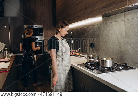 Smiling Young Woman In Apron Stand In Modern Kitchen Preparing Salad Use Smartphone Read Recipe, Hap