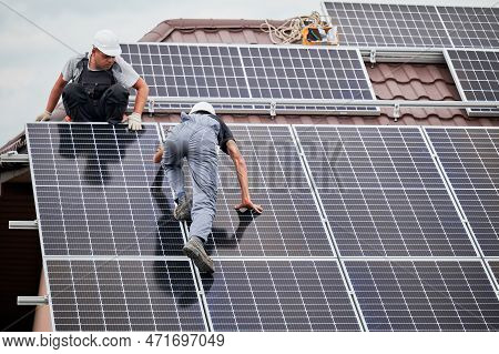 Men Mounters Mounting Photovoltaic Solar Moduls On Roof Of House. Electricians In Helmets Installing