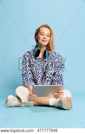 Cute Smiling Young Girl, Student Sitting On Floor With Tablet Over Blue Studio Background. Education
