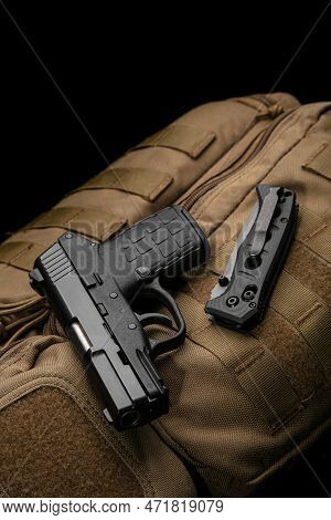 A Small Modern Pistol And Penknife On A Brown Military Backpack. Self Defense Kit. Dark Background.