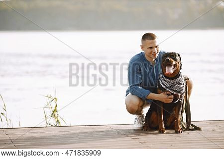 Man With Rotweiller Dog Sitting Together Near The Lake