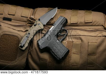 A Small Modern Pistol And Penknife On A Brown Military Backpack. Self Defense Kit. Dark Background.