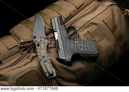 A Small Modern Pistol And Penknife On A Brown Military Backpack. Self Defense Kit. Dark Background.