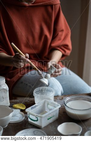 Female Ceramicist Or Potter Holding Paintbrush Applying Glaze On Ceramic Pieces While Sitting At Wor