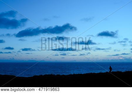 The Silhouette Of A Man Taking A Walk At Twilight, As The Sea And Sky Turn A Dramatic Deep Indigo Bl