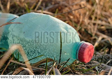 Crumpled Plastic Bottle Covered With Ice And Snow In Frozen Dry Grass. Green With Red Bottle Cap. Pl
