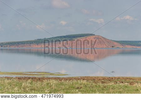 Lake With A Red Hill With Forest In The Background.gray Heron Flies And Is Reflected In The Lake.