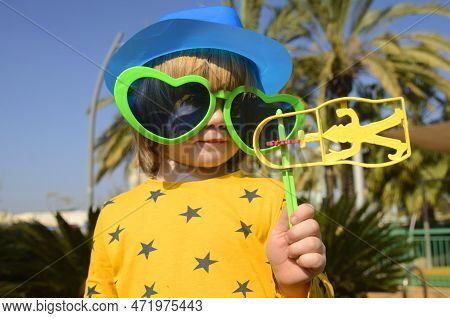 Carnival, Purim In Israel. A Cheerful Little Boy In A Hat, Funny Glasses, A Wig With A Rattle, Raash