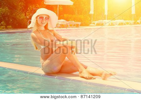 Beautiful female in white bikini seating near the waterpool under sun light