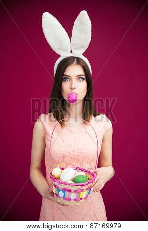 Woman in bunny ears holding Easter egg basket over pink background. Looking at camera. Holding egg with her lips. 