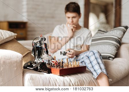 Play chess. Clever teenager putting his robot near chess board holding fingers near mouth while thinking about next move